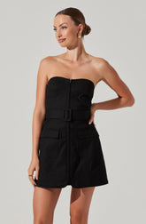 A woman is wearing a strapless black romper with a front zipper and a wide belt with a rectangular buckle. The romper has large front pockets on each side. She has her hair styled in a low bun and is accessorizing with gold earrings and a ring.